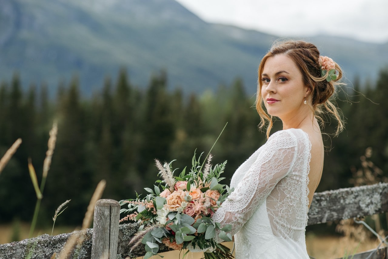 A bride holding her bouquet leans against a rustic fence.