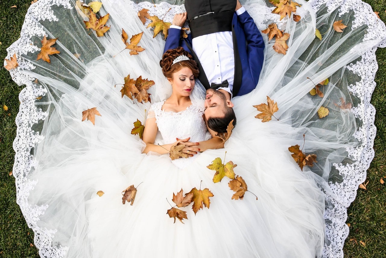 A bride and groom lay down on the ground as leaves fall on them.