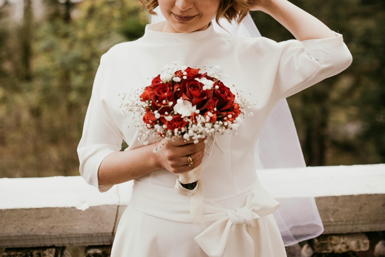 A bride wearing a ¾ length sleeve wedding gown and holding a bouquet of roses in an autumn forest.