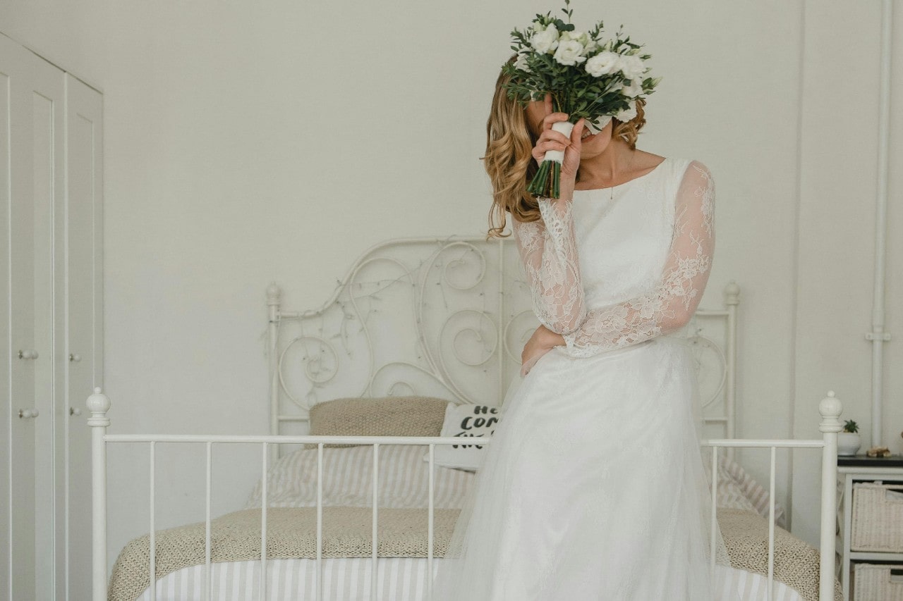 A bride holding her bouquet up to her face and resting against a white bed frame