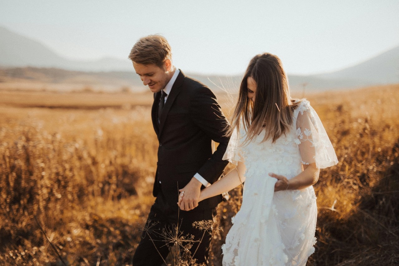 A bride and groom walking through a golden field during a clear fall day.