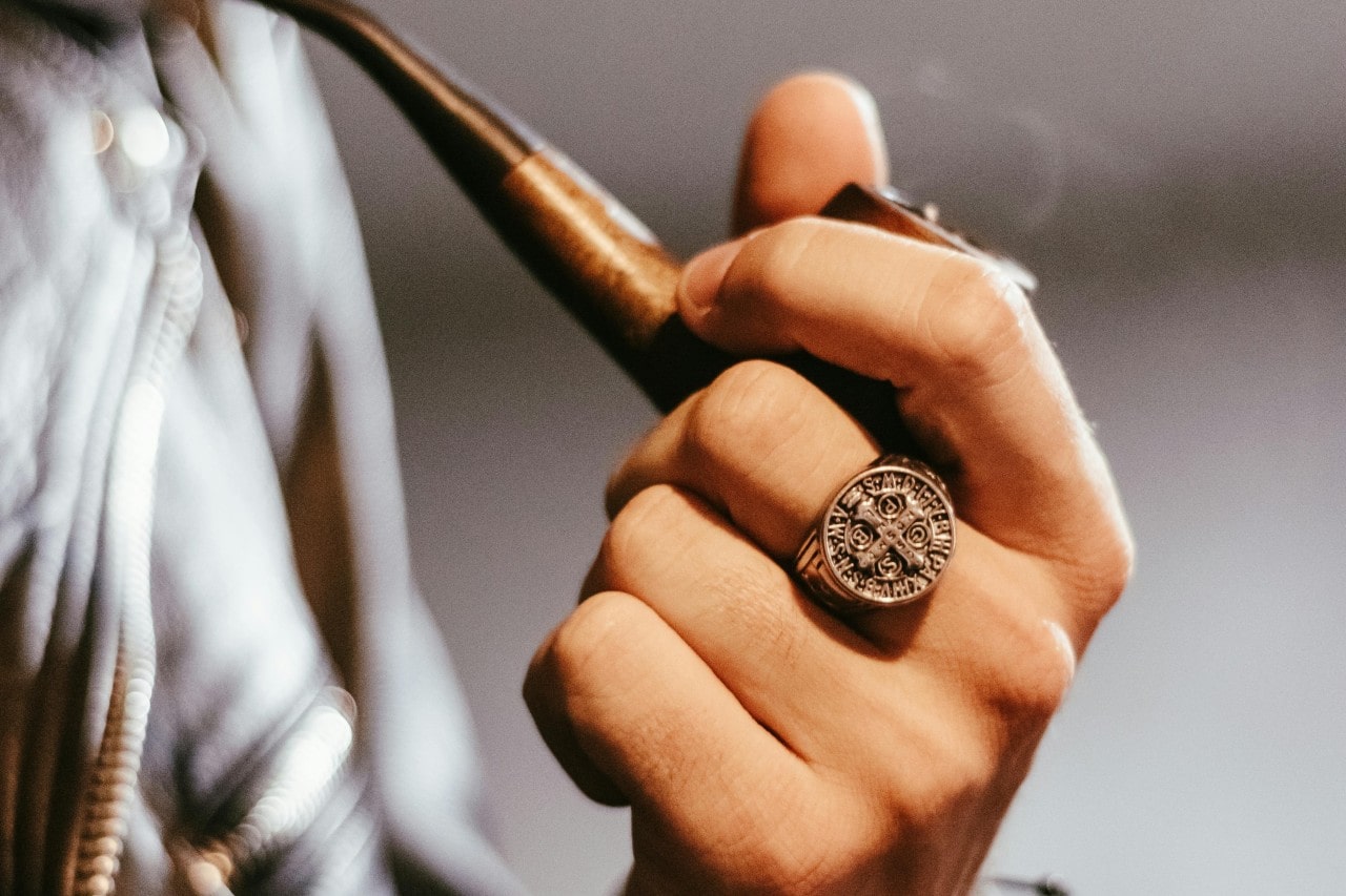 a person’s hand holding a pipe and wearing a cross signet ring