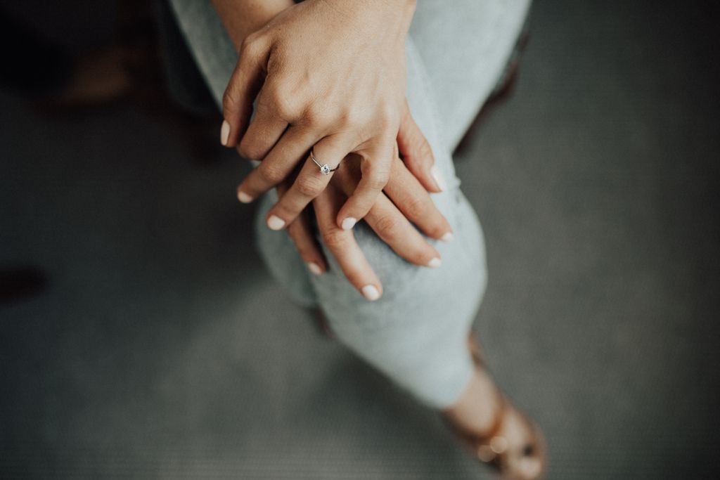 A woman sits with her legs crossed, a solitaire engagement ring on her finger.