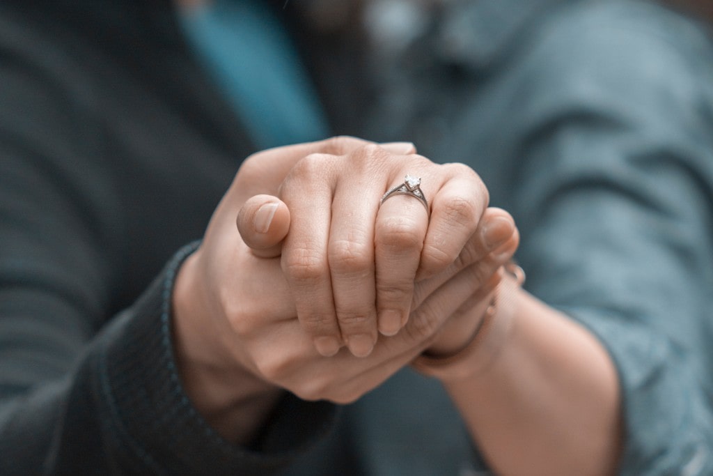 A couple holding hands, the engagement ring with a cathedral setting in clear view on her finger.