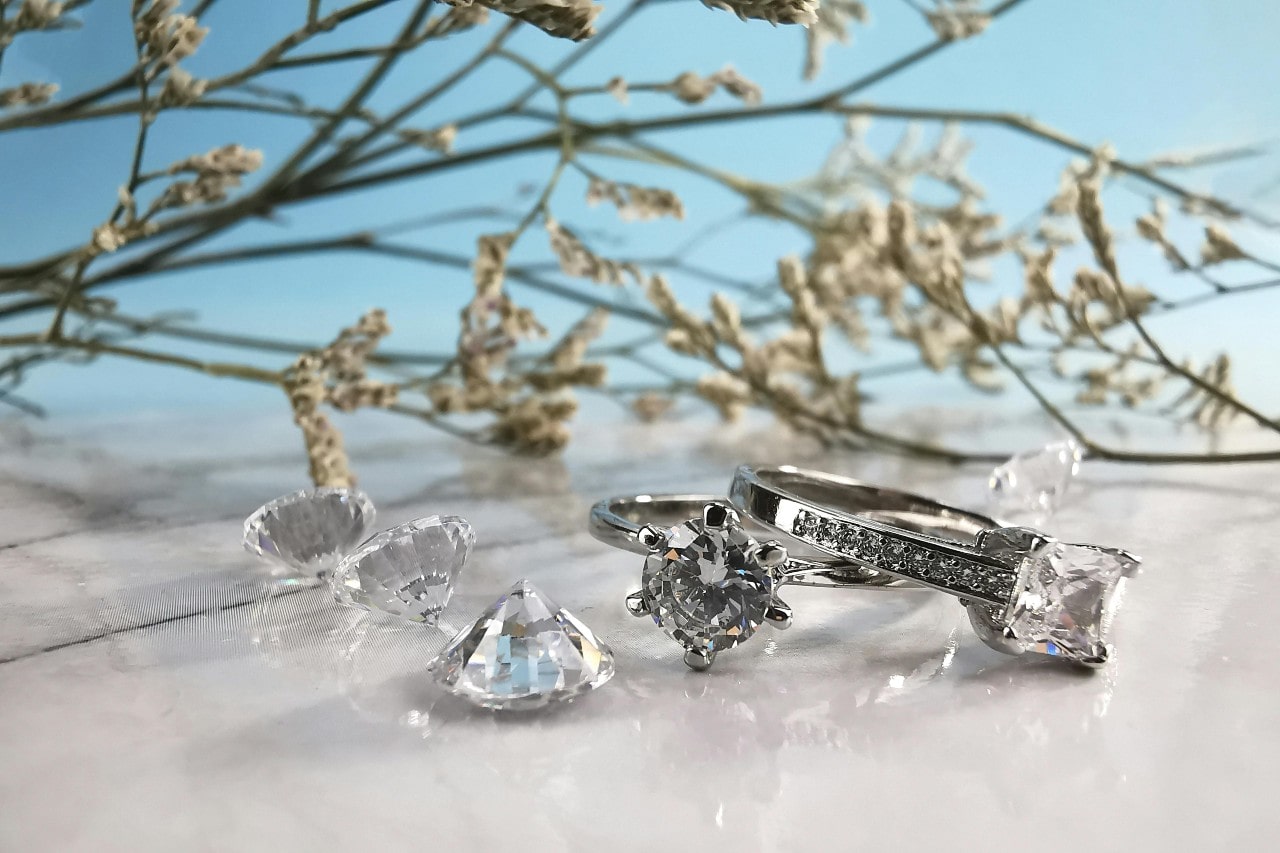 A close-up of two stunning diamond engagement rings displayed next to three round cut diamonds on a beachy background.