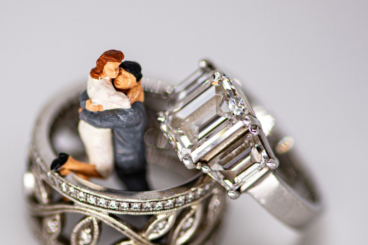 A close-up of a three stone engagement ring displayed next to two wedding bands around a small wedding figurine.